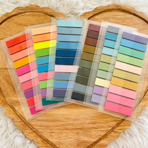 May include: A collection of colorful sticky note pads arranged on a wooden heart-shaped tray. Each pad features a gradient of colors, including pinks, oranges, greens, blues, and browns. The pads are in clear plastic packaging.
