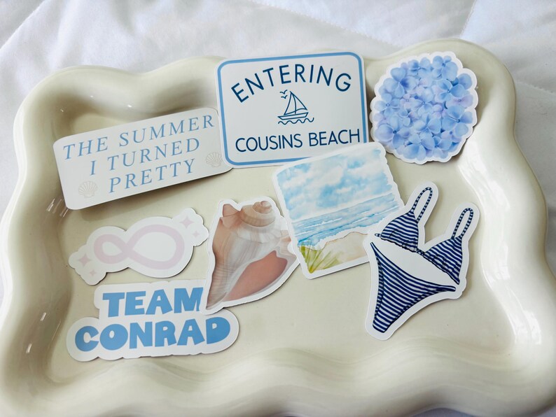 May include: A collection of stickers with a summer theme, including text that reads "The Summer I Turned Pretty", "Entering Cousins Beach", and "Team Conrad". Other stickers feature a seashell, a blue hydrangea, a bikini, and an infinity symbol.