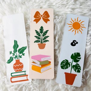 May include: Three bookmarks with different designs. The first bookmark is white with a green plant in a brown vase and a stack of books. The second bookmark is peach with a brown bow, a green plant in a brown pot, and a stack of books. The third bookmark is white with a yellow sun, a black ampersand, and a green plant in a brown pot.