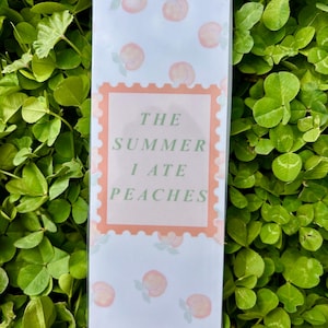 K&ouml;nnte beinhalten: Ein rechteckiger, hellfarbiger Gegenstand mit einem Pfirsichmuster und dem Text "THE SUMMER I ATE PEACHES" in einem pfirsichfarbenen Stempeldesign. Der Hintergrund ist ein Feld aus gr&uuml;nem Klee.