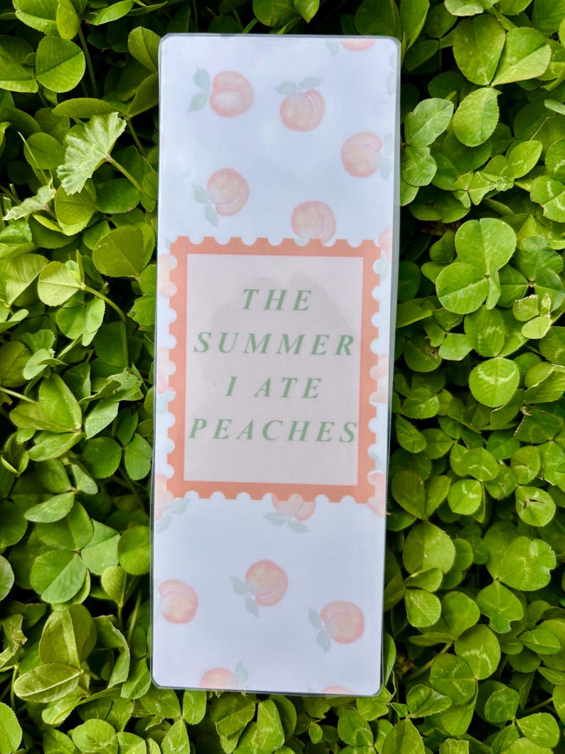 K&ouml;nnte beinhalten: Ein rechteckiger, hellfarbiger Gegenstand mit einem Pfirsichmuster und dem Text "THE SUMMER I ATE PEACHES" in einem pfirsichfarbenen Stempeldesign. Der Hintergrund ist ein Feld aus gr&uuml;nem Klee.