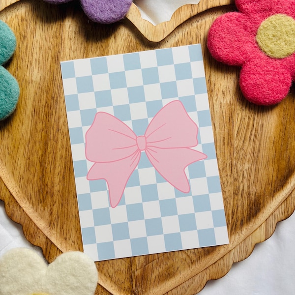 Peut inclure: Une carte à carreaux bleus et blancs avec un nœud rose au centre. La carte est sur un plateau en bois en forme de cœur avec des fleurs en feutre autour.