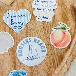 May include: A collection of stickers on a wooden tray. The stickers include a heart with "Connie Baby" and an infinity symbol, a circle with "Cousins Beach" and a sailboat, a peach, and a quote that says "He gave me the moon and the stars. Infinity."