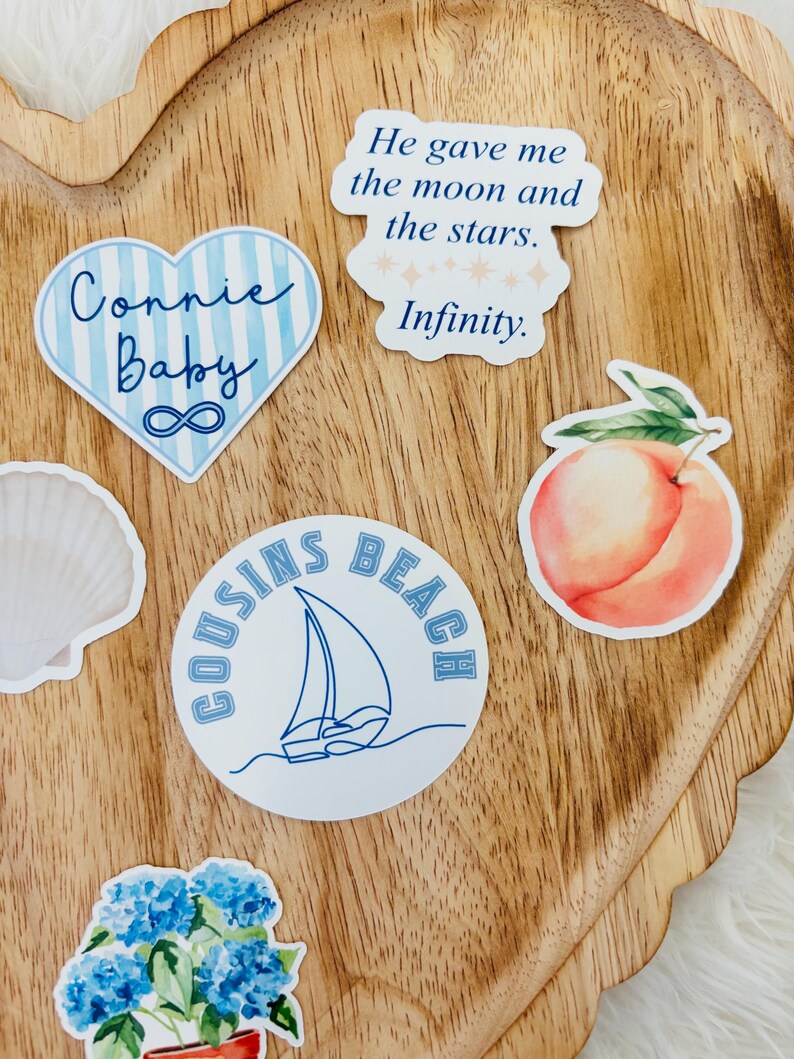 May include: A collection of stickers on a wooden tray. The stickers include a heart with "Connie Baby" and an infinity symbol, a circle with "Cousins Beach" and a sailboat, a peach, and a quote that says "He gave me the moon and the stars. Infinity."