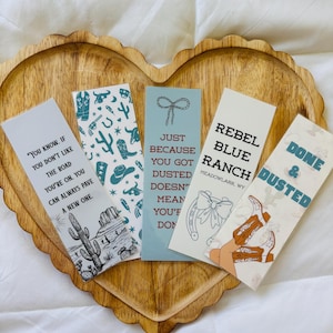 May include: Five rectangular bookmarks with western-themed designs and quotes are arranged on a wooden heart-shaped tray. The bookmarks feature illustrations of cacti, cowboy boots, and horseshoes, with phrases like "Rebel Blue Ranch" and "Done & Dusted."
