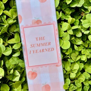 K&ouml;nnte beinhalten: Ein rechteckiger Lesezeichen mit einem rosa-wei&szlig;en Vichy-Muster und Pfirsich-Illustrationen. Der Text "THE SUMMER I YEARNED" ist in einem rosa Rahmen zentriert. Das Lesezeichen befindet sich vor einem Hintergrund aus gr&uuml;nem Klee.