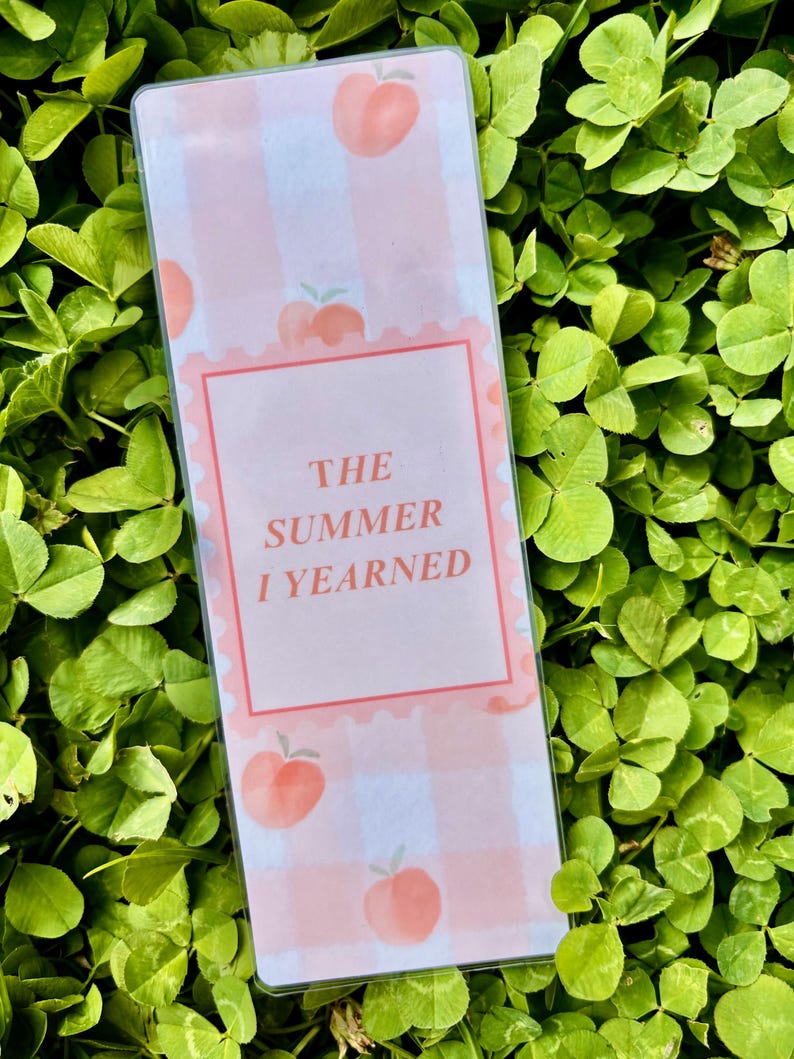 K&ouml;nnte beinhalten: Ein rechteckiger Lesezeichen mit einem rosa-wei&szlig;en Vichy-Muster und Pfirsich-Illustrationen. Der Text "THE SUMMER I YEARNED" ist in einem rosa Rahmen zentriert. Das Lesezeichen befindet sich vor einem Hintergrund aus gr&uuml;nem Klee.