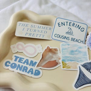 May include: A collection of stickers on a beige tray. The stickers include text that says "The Summer I Turned Pretty", "Entering Cousins Beach", and "Team Conrad". Other stickers feature a seashell, an infinity symbol, and a watercolor beach scene.