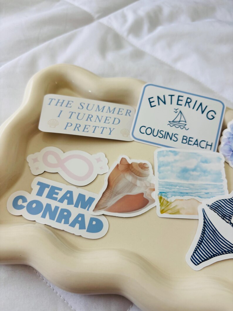 May include: A collection of stickers on a beige tray. The stickers include text that says "The Summer I Turned Pretty", "Entering Cousins Beach", and "Team Conrad". Other stickers feature a seashell, an infinity symbol, and a watercolor beach scene.