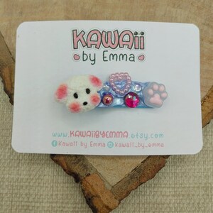 Peut inclure: Barrette Kawaii avec un chien blanc et des accents roses, un cœur rose et une empreinte de patte. La barrette est ornée d'ornements roses et bleus et du texte "KAWAII by Emma".