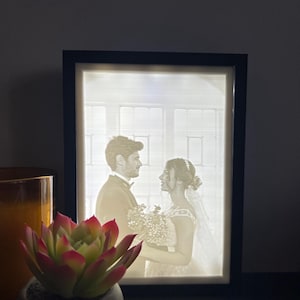 Könnte beinhalten: Ein beleuchteter 3D-gedruckter Bilderrahmen mit schwarzem Rahmen und weißem Hintergrund. Das Bild zeigt ein Paar in einer Hochzeitsszene.