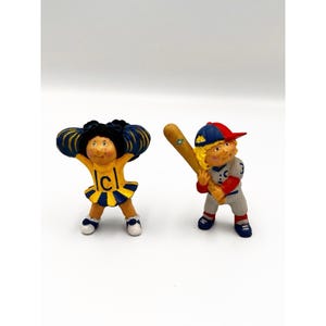 May include: Two small figurines: a cheerleader with dark hair wearing a yellow and blue uniform with a letter C, and a baseball player with blonde hair wearing a red and blue cap, a white pinstriped uniform, and holding a wooden baseball bat.