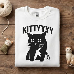 Könnte beinhalten: Weißes Sweatshirt mit dem Wort "KITTYYYY" über einer schwarzen Katzenzeichnung. Die Katze hat große Augen und Schnurrhaare. Das Sweatshirt liegt auf einer Holzoberfläche mit Katzen-Dekorationen.