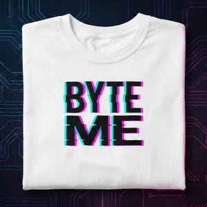 May include: White t-shirt featuring the text "BYTE ME" in a glitch effect. The text is black with cyan and magenta accents. The shirt is folded, set against a dark background.