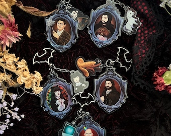 May include: Five colourful keychains featuring portraits of different people in ornate frames. Each keychain has a bat-shaped charm attached to the top.
