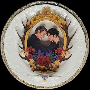 Puede incluir: Un plato decorativo blanco con borde dorado, con una imagen de dos hombres en un marco dorado, decorado con cornamentas y flores. El plato tiene un patrón en relieve. La imagen representa una escena romántica.