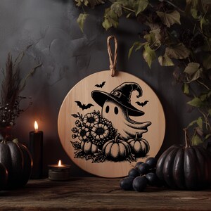May include: A round wooden Halloween decoration featuring a ghost wearing a witch hat, surrounded by pumpkins, flowers, and bats. The design is in black and white, with a natural wood background and a jute rope for hanging.