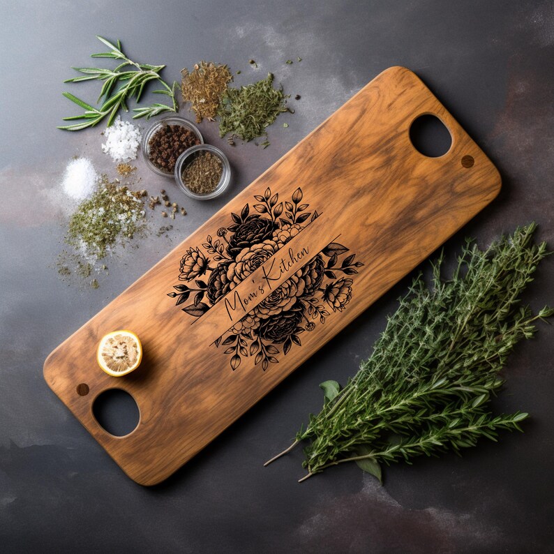 May include: A wooden serving board with a floral design and the text "Mom's Kitchen" engraved on it. The board has two handles and is made of acacia wood.