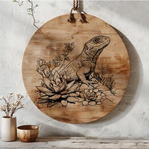 May include: A round wooden wall decoration featuring a black and white line drawing of a lizard sitting on a rock with succulents. The lizard is facing to the right and has a long tail.