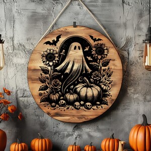 May include: Round wooden Halloween sign featuring a ghost, pumpkins, sunflowers, bats, and skulls. The design is etched in black on a light brown wood background. A rope is attached for hanging. The sign is displayed with orange pumpkins.