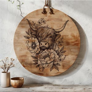 May include: Round wooden wall art featuring a detailed engraving of a Highland cow with large horns, surrounded by floral elements. The artwork is in shades of brown and tan, with a natural wood grain background and a rope for hanging.