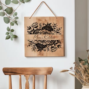 May include: A wooden sign with a floral design and the text "Mom's Kitchen" in black lettering. The sign is hanging from a rope and is in front of a white wall.