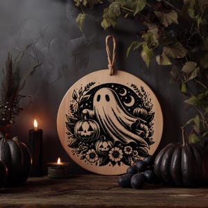 May include: Round wooden Halloween decoration featuring a ghost, pumpkins, flowers, and crescent moons. The design is etched in black against the natural wood grain. A jute loop is attached for hanging. The scene is set with lit candles and dark pumpkins.