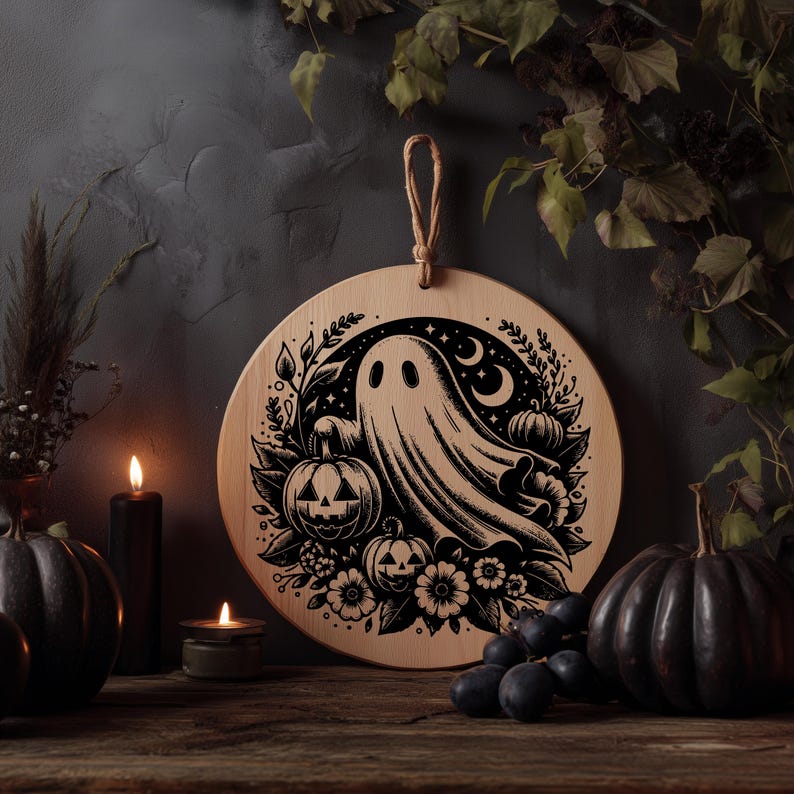 May include: Round wooden Halloween decoration featuring a ghost, pumpkins, flowers, and crescent moons. The design is etched in black against the natural wood grain. A jute loop is attached for hanging. The scene is set with lit candles and dark pumpkins.