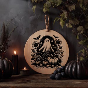 May include: A round wooden cutting board with a Halloween-themed design. The black illustration features a ghost, pumpkins, sunflowers, roses, bats, and skulls. A rope loop is attached for hanging. The board is displayed with black pumpkins, candles, and grapes.