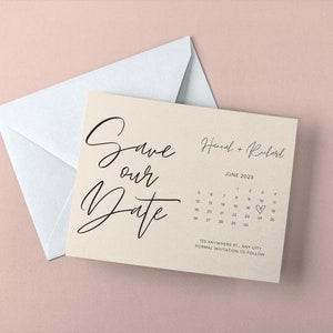 May include: A cream-colored 'Save the Date' card with black script and a calendar for June 2023. The card is paired with a white envelope, set against a soft pink background. Text includes names and address.