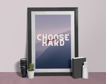 May include: A framed print with a motivational quote, "CHOOSE your HARD", against a backdrop of a hazy mountain range. The print is in a black frame and is displayed on a table with a camera lens, a potted plant, a book, and a bottle.
