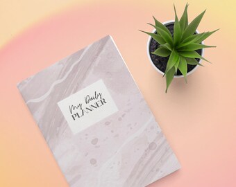 May include: A white planner with a marble pattern and the text "My Daily Planner" on the front cover. The planner is on a pink and yellow gradient background with a small green plant in a white pot.