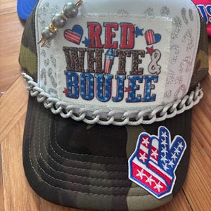 Peut inclure: Une casquette trucker camouflage avec un panneau avant blanc à motif léopard argenté pailleté. Un écusson indique RED WHITE & BOUJEE avec des motifs patriotiques. La casquette est ornée d'une chaîne argentée, d'une épingle perlée et d'un écusson main.