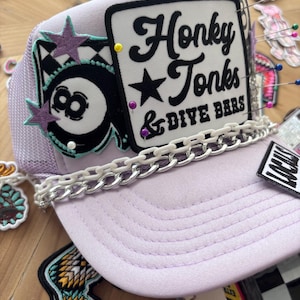 May include: A lavender trucker hat with a white chain detail and the words "Honky Tonks & Dive Bars" on a black and white sign. The hat features an 8-ball patch and star accents.