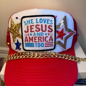 Pode incluir: Boné de caminhoneiro vermelho, branco e azul com o texto "She Loves Jesus and America Too". O boné apresenta estrelas douradas e com lantejoulas, uma corrente dourada e alfinetes decorativos.