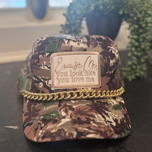 Pode incluir: Boné de caminhoneiro com estampa camuflada, detalhe de corrente dourada e um patch que diz "Excuse Me, you look like you love me". O boné tem uma parte traseira de malha e uma aba curva.