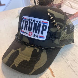 May include: A camouflage trucker hat with a black and white patch that says "TRUMP 47" with red, white, and blue stars and stripes. The hat has a black chain trim and a black and gold beaded embellishment.