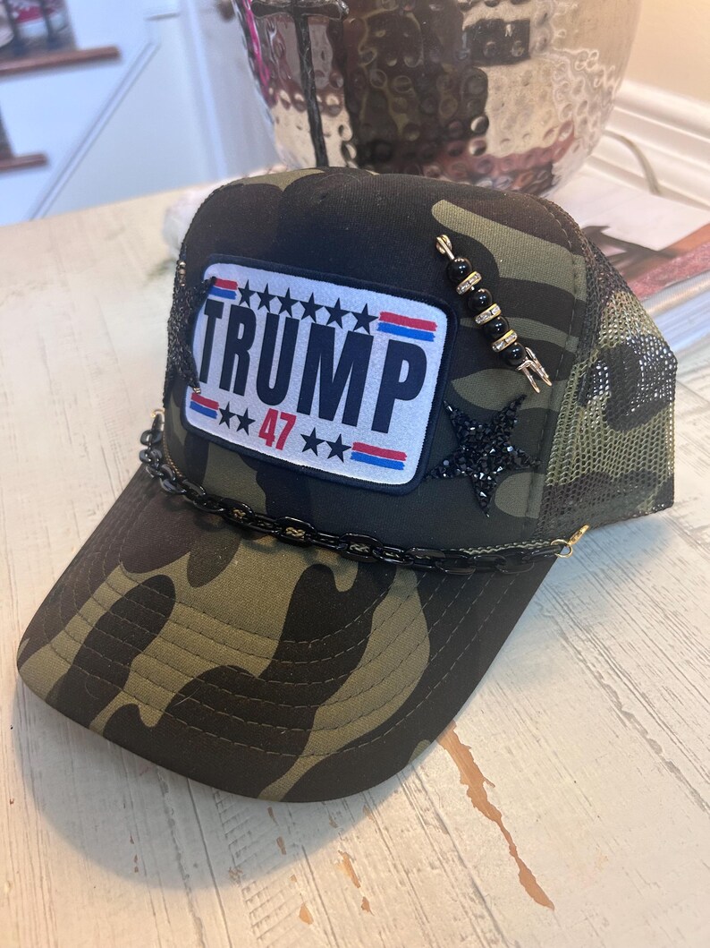 May include: A camouflage trucker hat with a black and white patch that says "TRUMP 47" with red, white, and blue stars and stripes. The hat has a black chain trim and a black and gold beaded embellishment.
