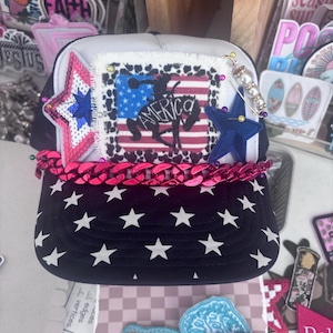 Pode incluir: Um boné de beisebol patriótico com costas de malha branca e aba azul marinho adornada com estrelas brancas. A frente apresenta um patch com um desenho da bandeira americana e a palavra "America". Corrente rosa e enfeites de estrela.