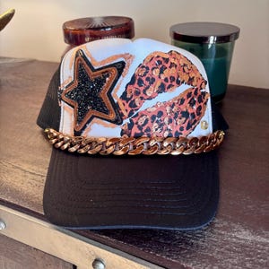 May include: A black and white trucker hat with a gold chain, a star with black glitter, and leopard print lips. The hat has a black brim and mesh back.