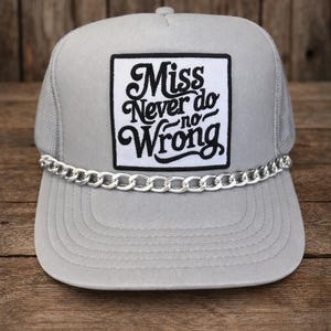 Pode incluir: Boné cinza de caminhoneiro com detalhe de corrente prateada e um patch quadrado com a frase "Miss Never do no Wrong" em escrita preta. O boné tem uma parte traseira de malha e uma viseira curva. Sobre uma superfície de madeira.