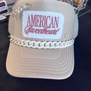 Pode incluir: Boné de caminhoneiro bege com detalhe de corrente branca na aba. O boné apresenta o texto "AMERICAN Sweetheart" em escrita vermelha em um patch branco. Um alfinete de pérolas e ouro e um pequeno strass também estão anexados.
