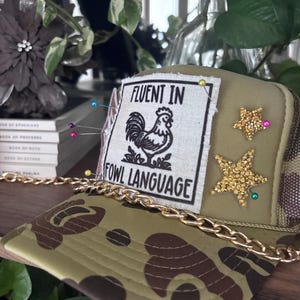 Pode incluir: Um boné de caminhoneiro camuflado com uma corrente dourada e um patch que diz "FLUENT IN FOWL LANGUAGE" com um gráfico de galo. O chapéu é decorado com enfeites dourados em forma de estrela e alfinetes coloridos.