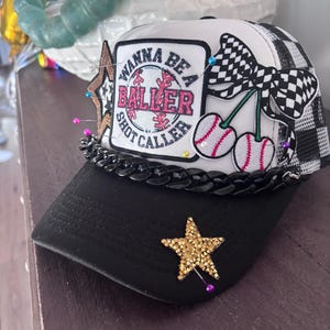 May include: A black and white baseball cap with the text "Wanna Be a Baller Shot Caller" on a patch. The cap features a gold star, a checkered bow, and cherry designs. The brim is black.