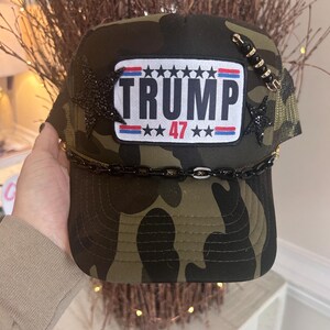 May include: A camouflage trucker hat with a black patch that says "TRUMP 47" in white letters. The patch has a red, white, and blue American flag design. The hat has black stars and a gold chain around the brim.