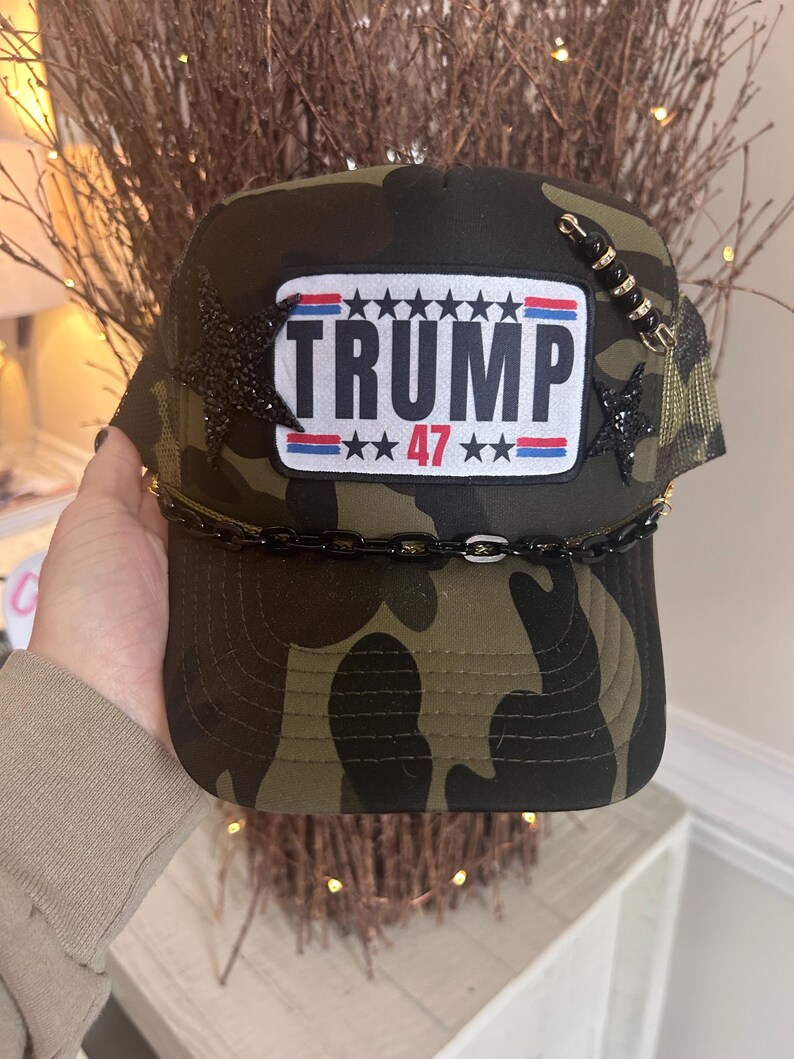 May include: A camouflage trucker hat with a black patch that says "TRUMP 47" in white letters. The patch has a red, white, and blue American flag design. The hat has black stars and a gold chain around the brim.