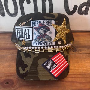 Pode incluir: Boné de caminhoneiro camuflado com costas em malha, adornado com patches e enfeites. Apresenta um patch com um gráfico de cowgirl e o texto "BORN FREE BUT NOW I'M EXPENSIVE". Inclui uma estrela dourada, um pequeno patch da bandeira americana e uma corrente.