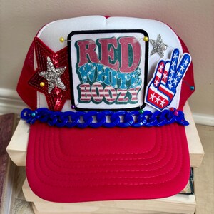 Pode incluir: Boné de caminhoneiro vermelho, branco e azul com o texto "RED WHITE BOOZY" em um patch preto. O boné apresenta uma aba vermelha, uma frente branca e uma parte traseira de malha vermelha. Enfeites incluem uma estrela e um sinal de paz.