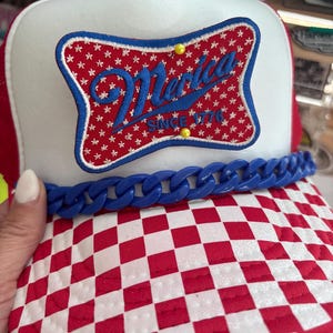 Pode incluir: Boné de caminhoneiro vermelho, branco e azul com viseira xadrez. O boné apresenta um patch com a palavra "Merica" em escrita azul, rodeada por estrelas, e o texto "Since 1776". Um detalhe de corrente azul adorna a aba.