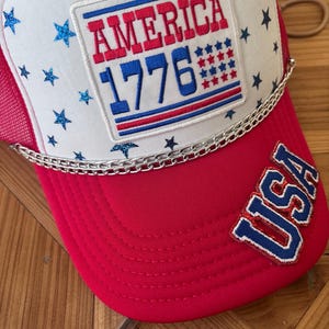 Pode incluir: Um boné de caminhoneiro vermelho, branco e azul com o texto "AMERICA 1776" na frente. O boné apresenta um painel frontal branco com estrelas brilhantes azuis, uma aba vermelha e um detalhe de corrente prateada. A palavra "USA" é bordada na lateral.